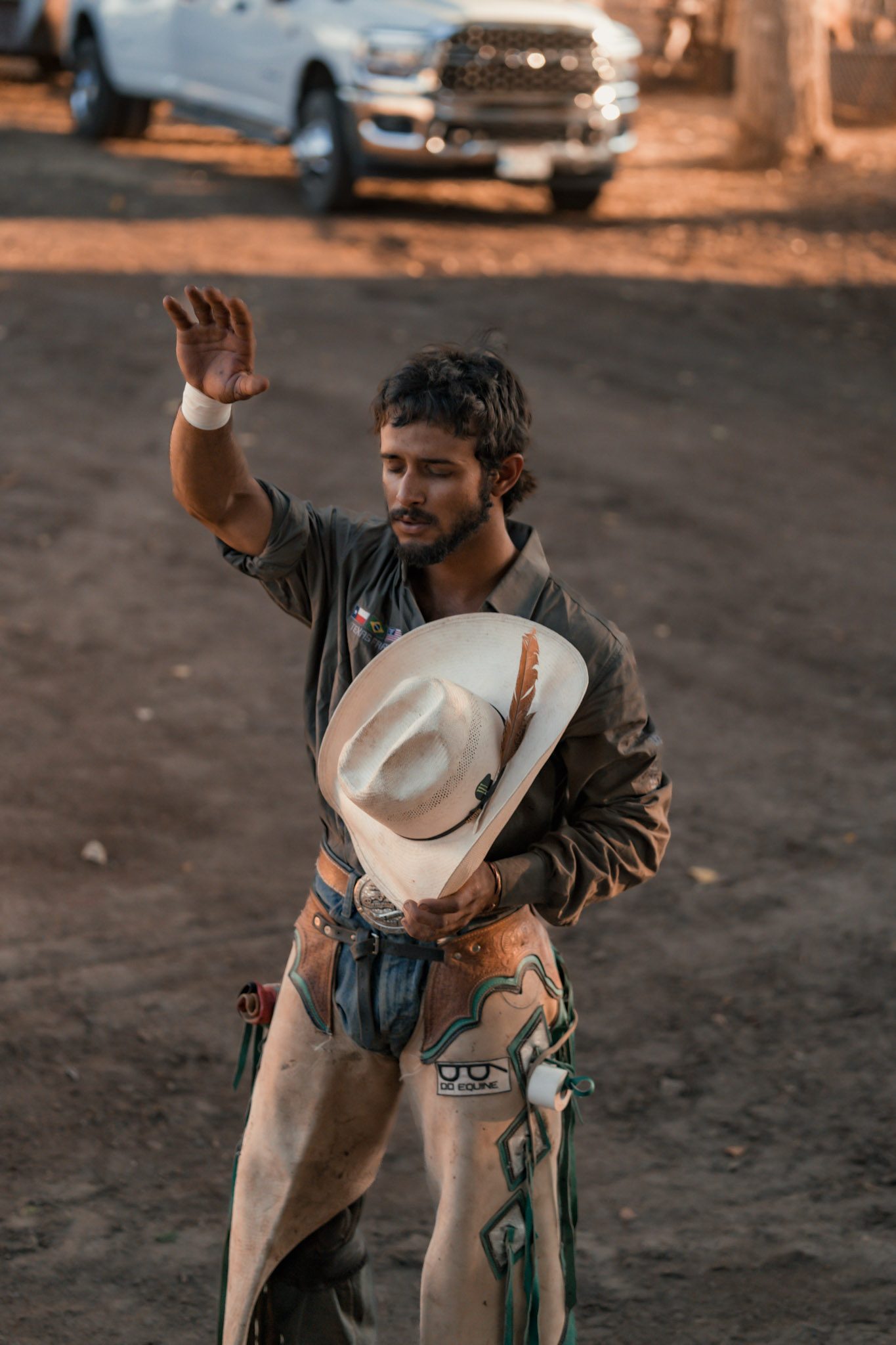 A cowboy in chaps holding his hat against his chest with one hand raised in worship