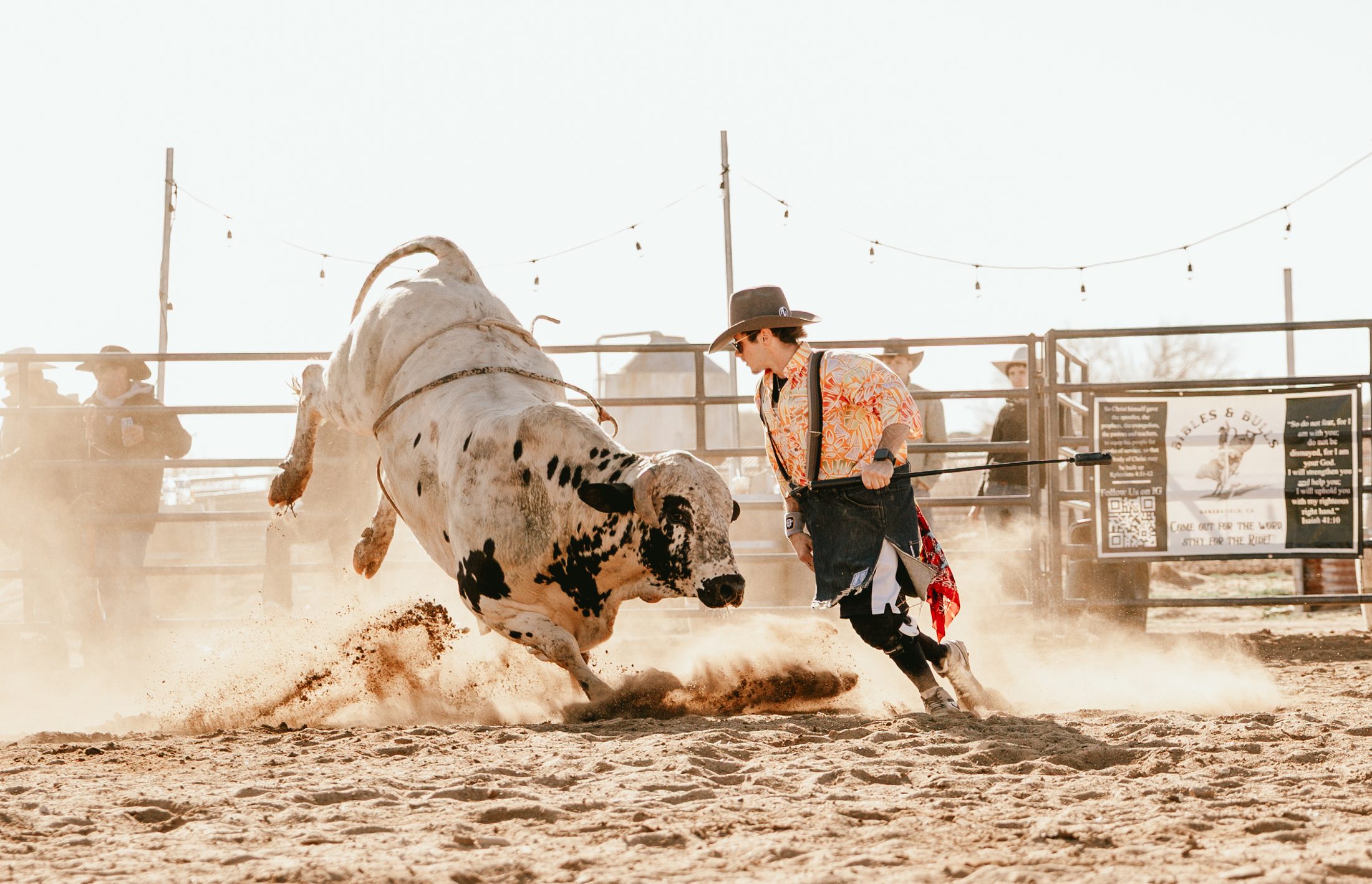 A bull bucking through the dirt as a bullfighter steps in, under string lights at a Bibles and Bulls gathering