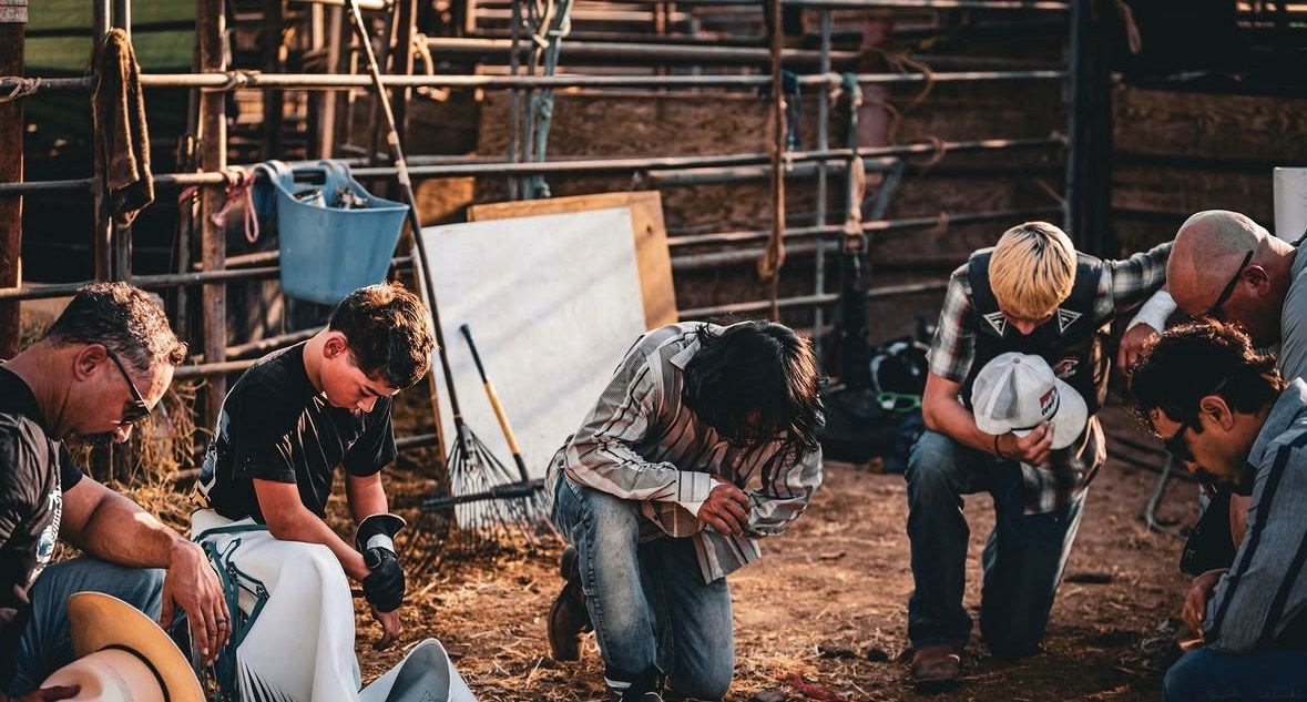 Riders and older men bowed together in prayer beside the chutes before an event